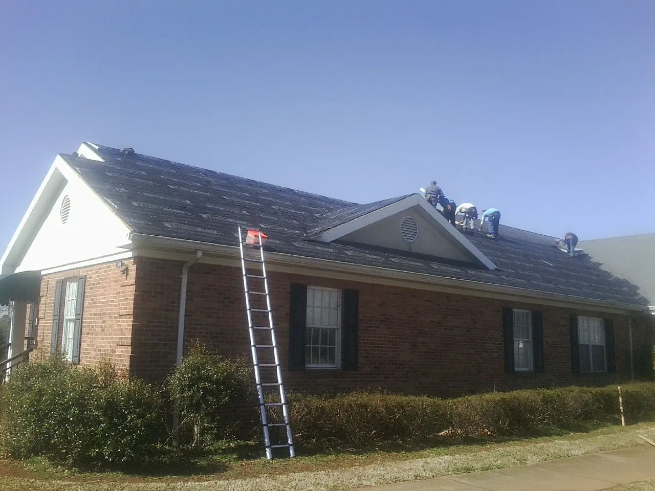 Crew tearing shingles off a brick ranch roof with a ladder set — Emergency Roof Repair work by our crew in Pine Prairie