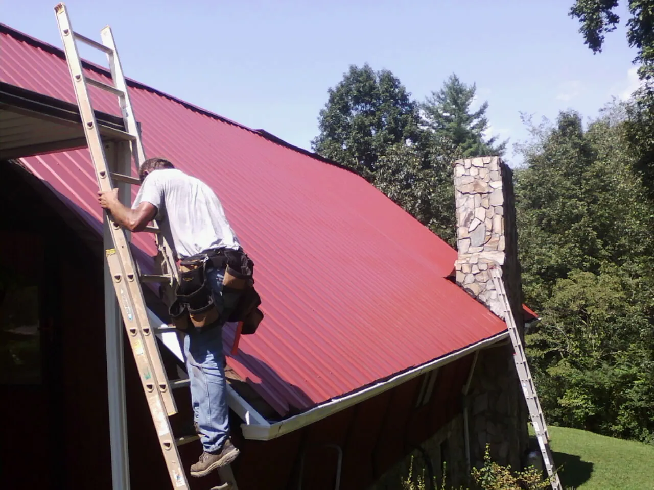 Roofer on a ladder beside a red metal roof and stone chimney — Soffit & Fascia Repair by Roofing Brothers in Pine Prairie